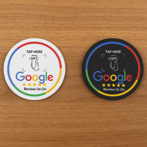 Two round NFC-enabled “Review Us On Google” coasters placed on a wooden surface — one white and one black — each featuring the Google logo, five yellow stars, and text instructing users to “TAP HERE” to leave a review.