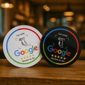 Two round NFC-enabled “Review Us On Google” tap-to-review signs, one white and one black, displayed on a wooden counter in a warm, blurred café setting.